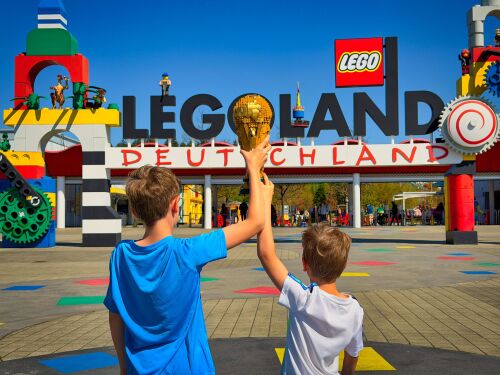 Samy und Tobi halten den offiziellen LEGO Pokal der FIFA Fußball-Weltmeisterschaft in die Höhe und freuen sich schon auf das neue Fußballevent FIFA World Cup 2026 Experience vom 11. Juni bis 19. Juli in den LEGOLAND Resorts.